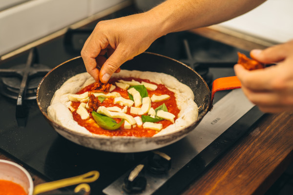 Hand putting toppings onto pizza dough in a frying pan